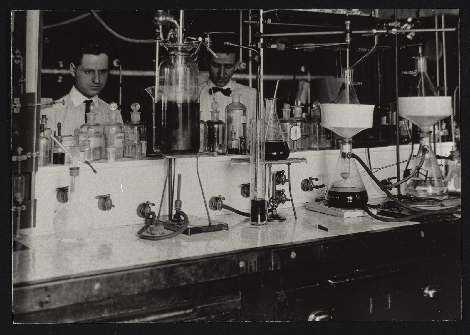 Two chemists behind a bench crowded with flasks, funnels and bottles in the Bureau of Chemistry Carbohydrate Laboratory, 1910s
