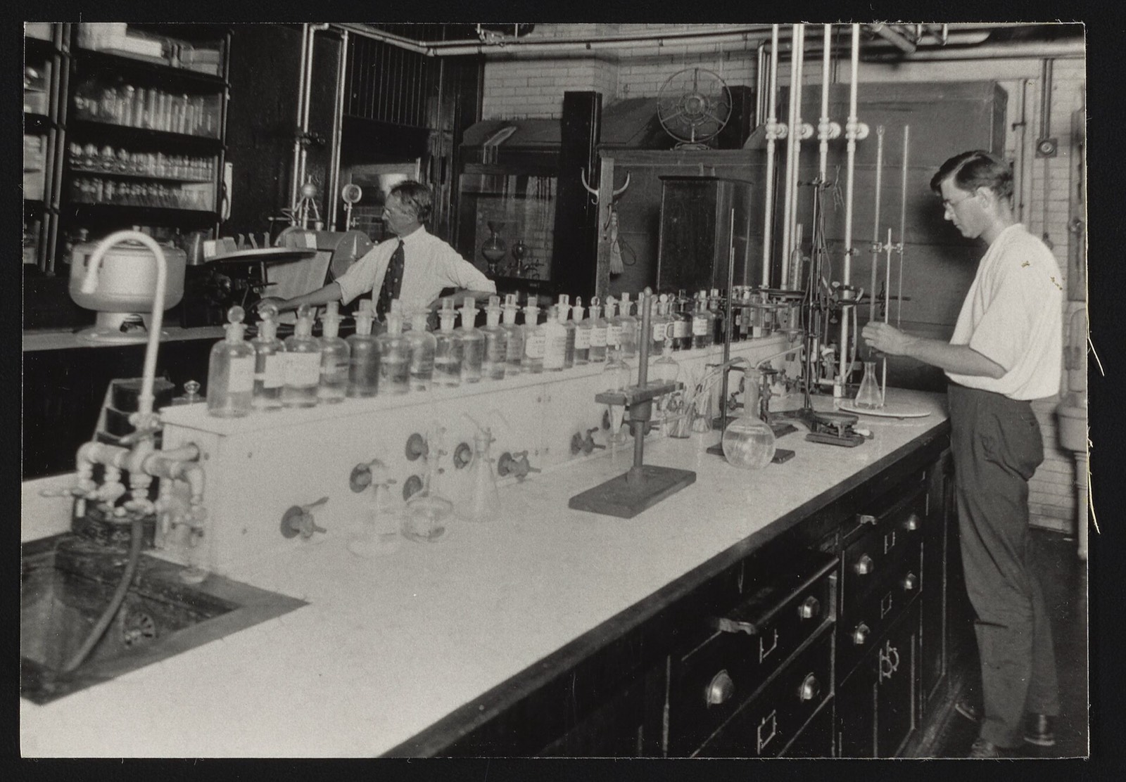 Two chemists at a long bench of bottles and burettes in the Bureau of Chemistry Drug Laboratory, 1900s–1920s