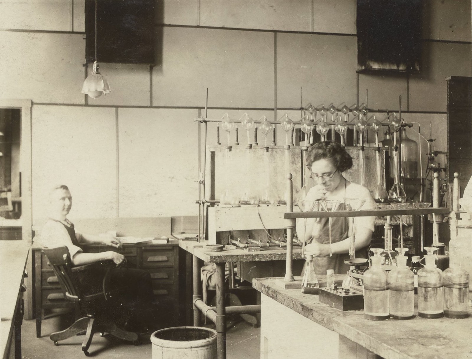 A small analytical laboratory with glassware on a bench; one researcher seated at a desk, another standing and blowing into a glass apparatus