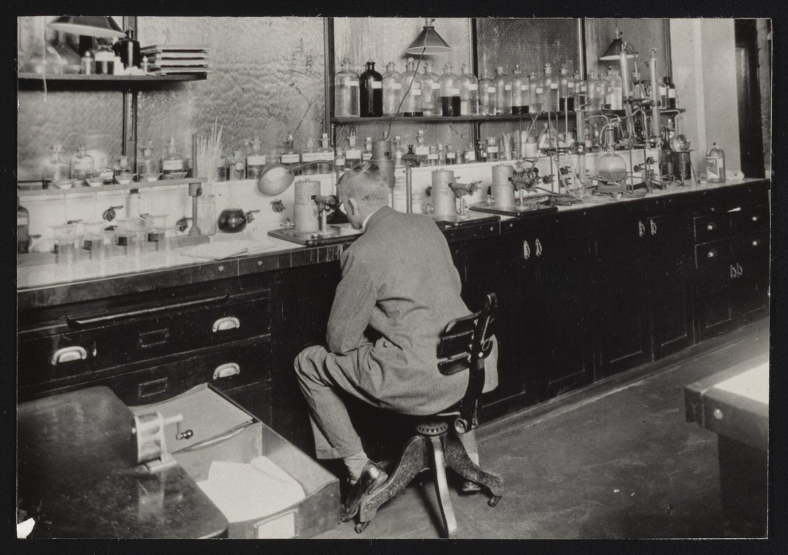 A chemist seated at a microscope with his back to the camera, before a wall of sample jars in the Bureau of Chemistry Water and Beverage Laboratory